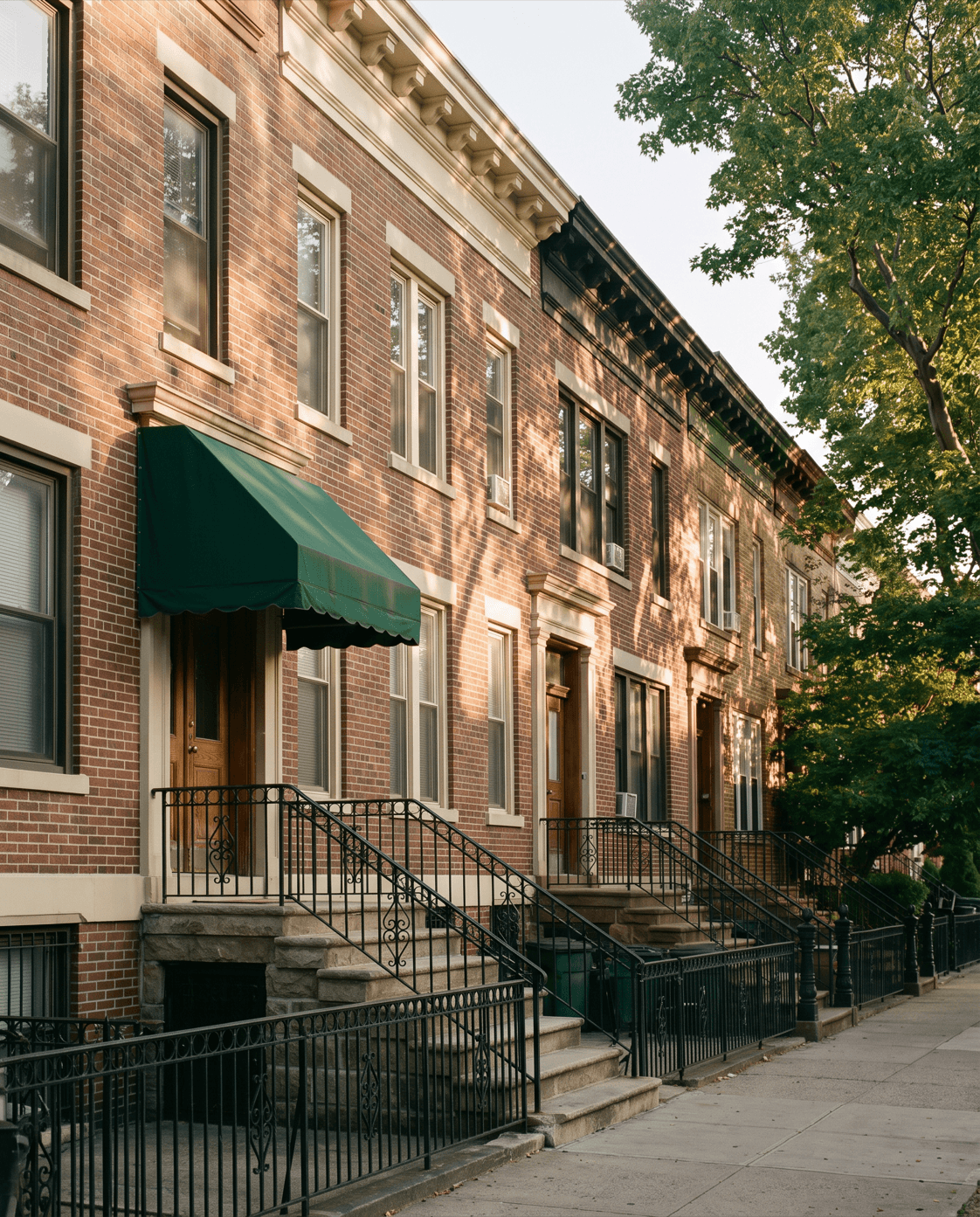 Row of brick Astoria rowhouses with stone stoops and a green awning in golden-hour light